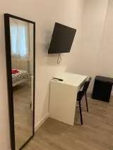 Televisor de pared sobre un escritorio con silla negra, espejo de cuerpo entero y minibar